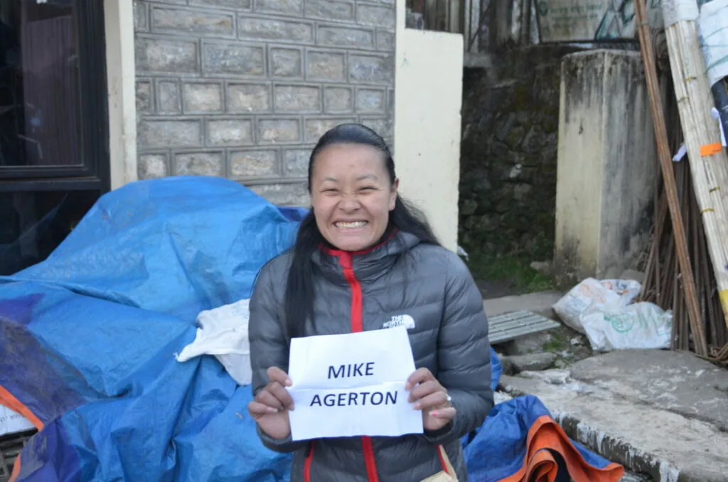 Woman smiling while holding a nameplate reading Mike Agerton in a cluttered alleyway with blue tarps and stored materials
