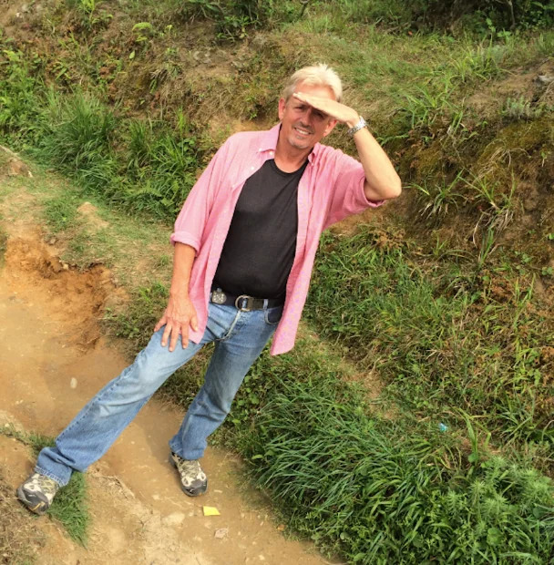 Man in pink shirt and straw hat posing on dirt path with grassy hillside behind him