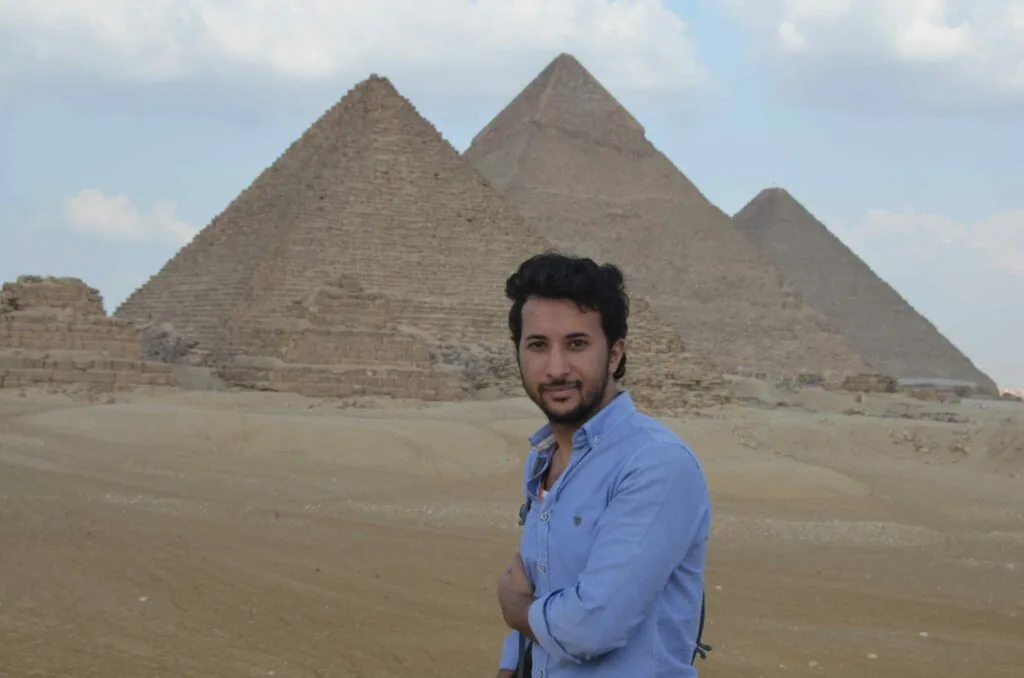 Man in blue shirt posing in front of ancient pyramids in desert landscape