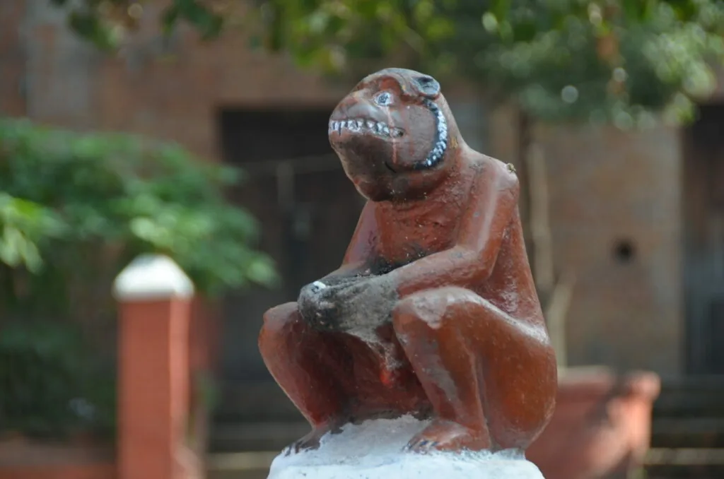 Brown wooden sculpture of a seated figure with distinctive facial features on a white base in a brick courtyard