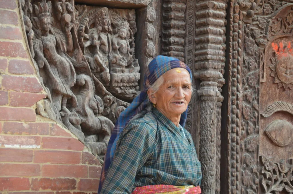 Elderly person in blue headwrap and checkered shirt standing against ornately carved wooden door or architectural element