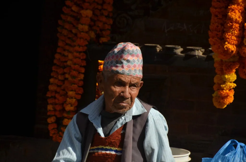 Elderly man wearing a patterned cap and traditional clothing sitting indoors with hanging marigolds and brick walls in background