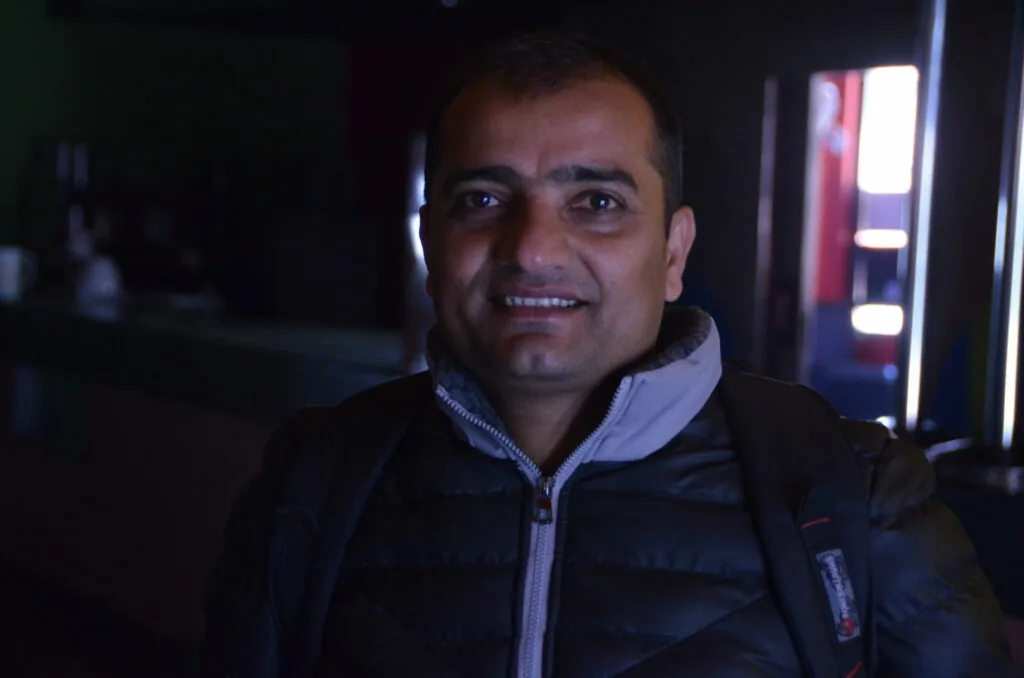 Man smiling at camera wearing dark jacket with blue collar in dimly lit indoor setting