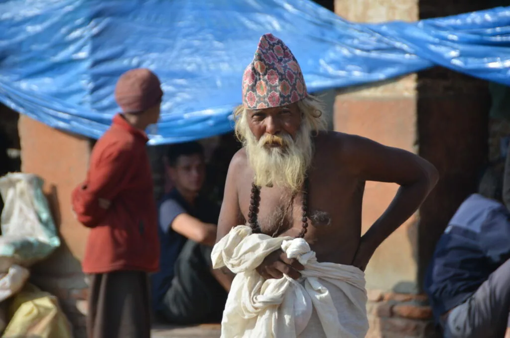 Elderly man with long white beard wearing traditional patterned cap and prayer beads, standing under blue tarp with other people in background