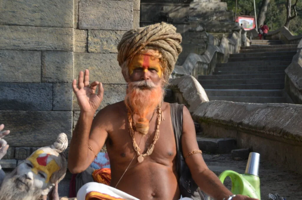 Shirtless man with long gray beard and wrapped turban waving, with religious markings on face, sitting at stone temple steps