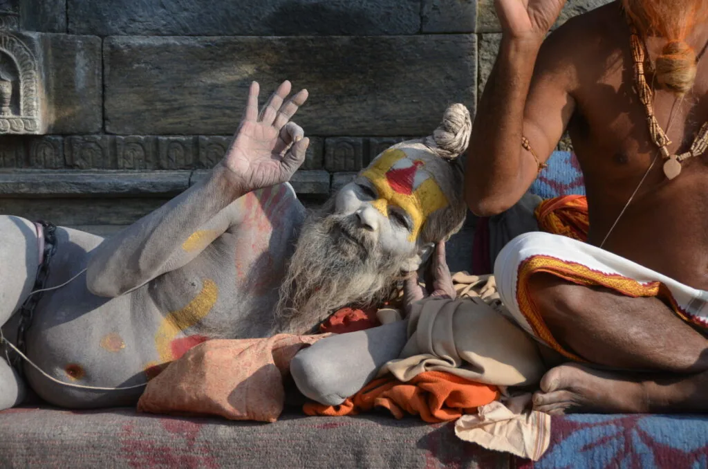Person with face and body painted in white, yellow and red sitting cross-legged against a stone wall