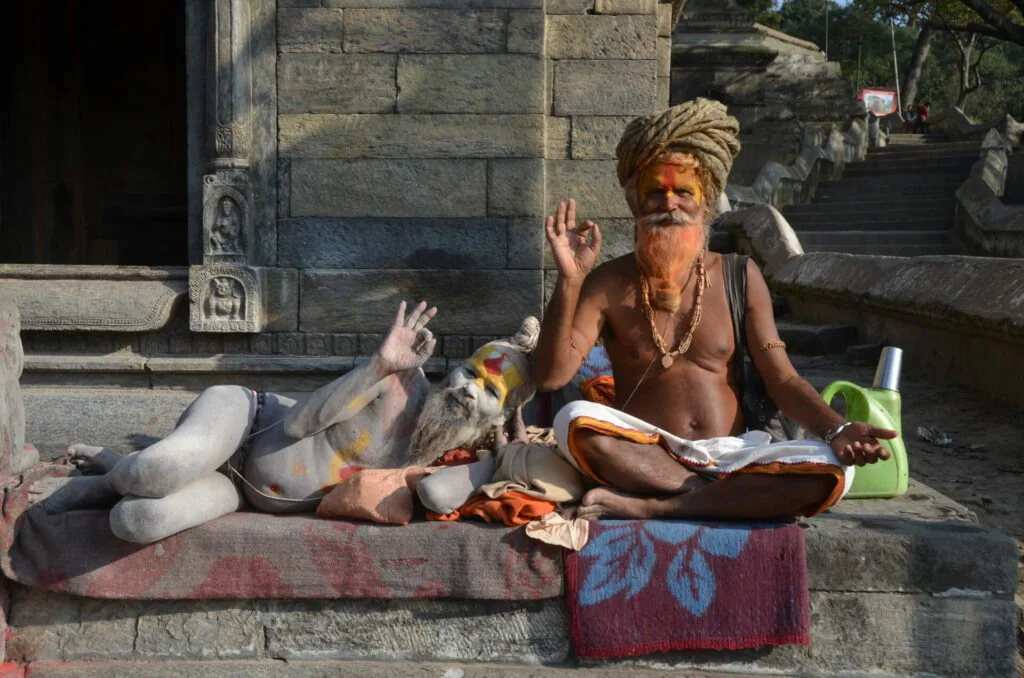 Elderly man with white beard and yellow turban sitting cross-legged on stone steps, holding religious text, with spiritual markings on body