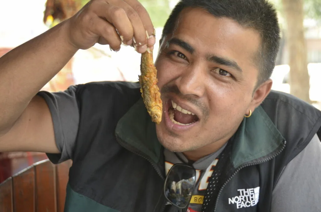Man in black jacket holding up a fried snack while smiling at camera