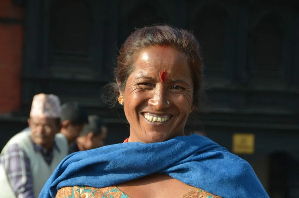 Woman wearing blue shawl with red tilaka mark on forehead, smiling at camera