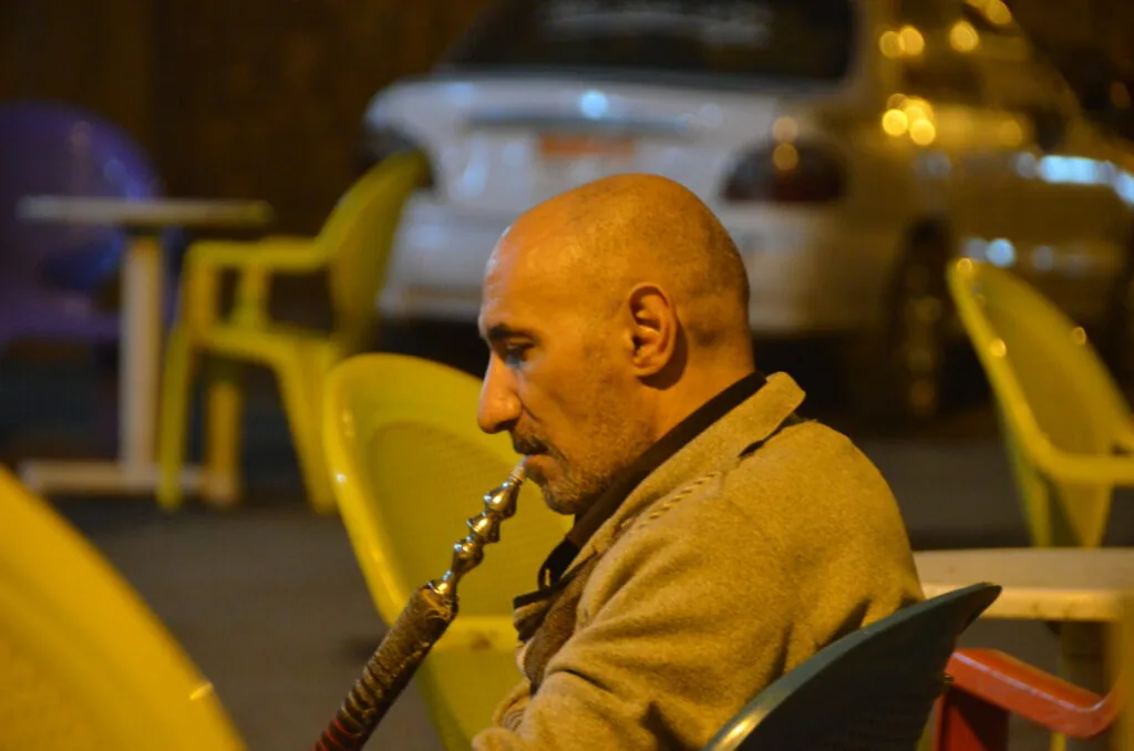 Man smoking hookah at outdoor café with yellow chairs and parked cars in background at night