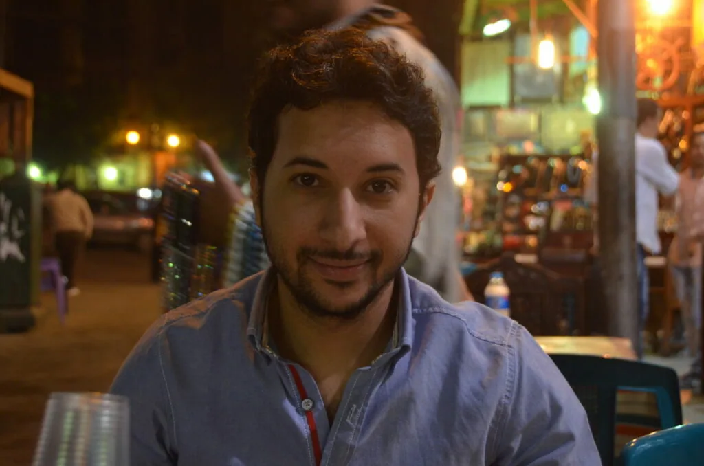 Man with dark hair and beard smiling at camera on illuminated nighttime street