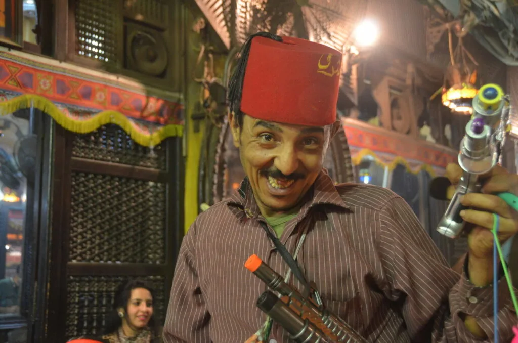 Man wearing red fez hat and pinstriped shirt holding musical instrument in decorated interior setting