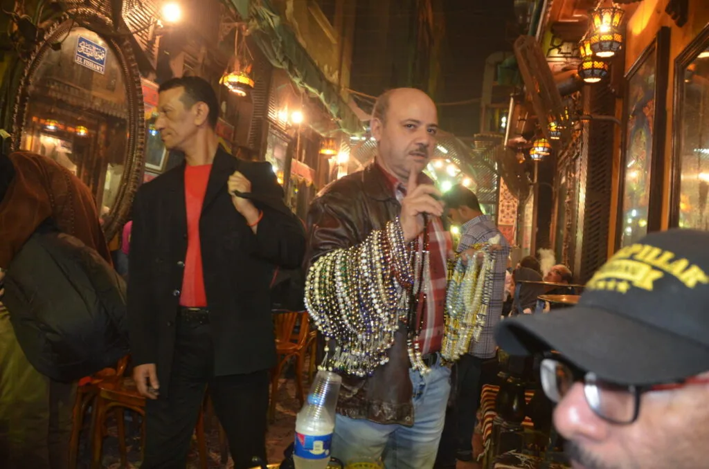 Man wearing colorful beaded necklaces holding microphone at nighttime street celebration with onlookers
