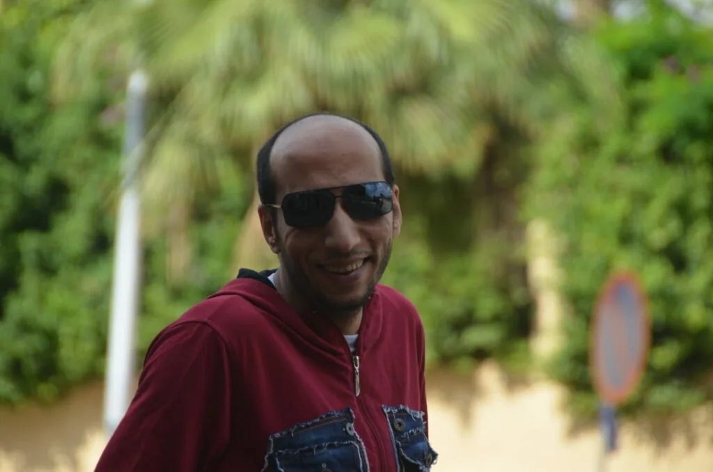 Man wearing dark sunglasses and maroon hoodie smiling at camera with green foliage in background