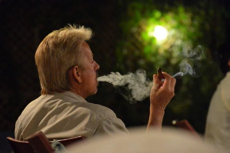 Man with light-colored hair smoking a cigar outdoors at night with blurred foliage and lighting in background