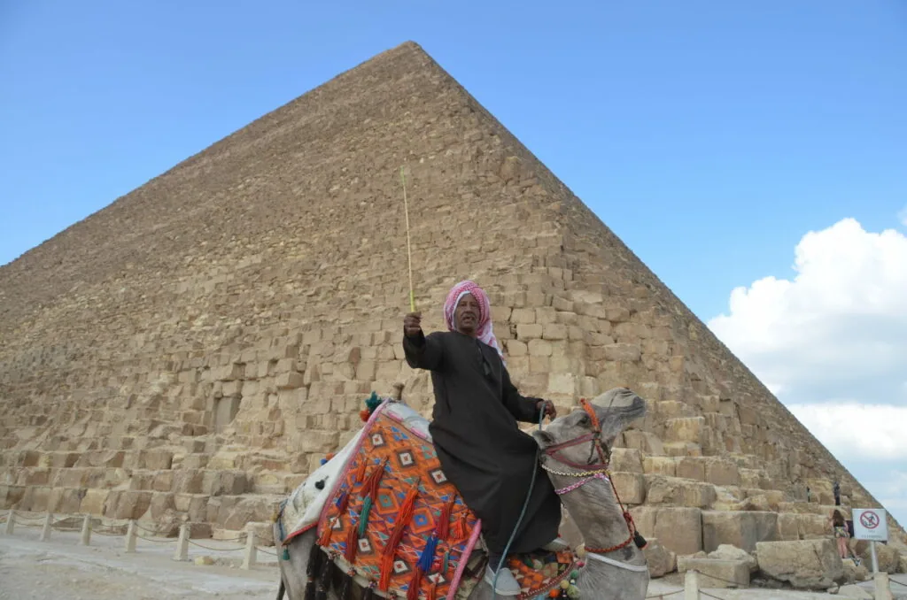 Person in pink headscarf and black clothing riding a camel with orange decorative blanket in front of a large stone pyramid under blue sky