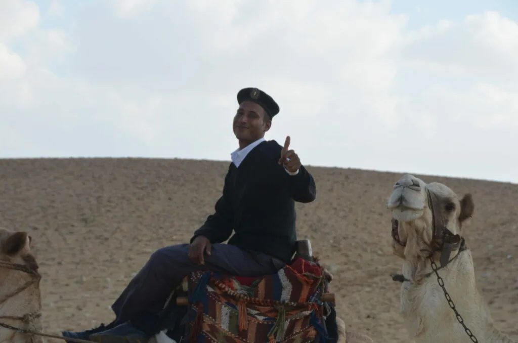 Man in business suit sitting on a camel in desert landscape, making a peace sign gesture