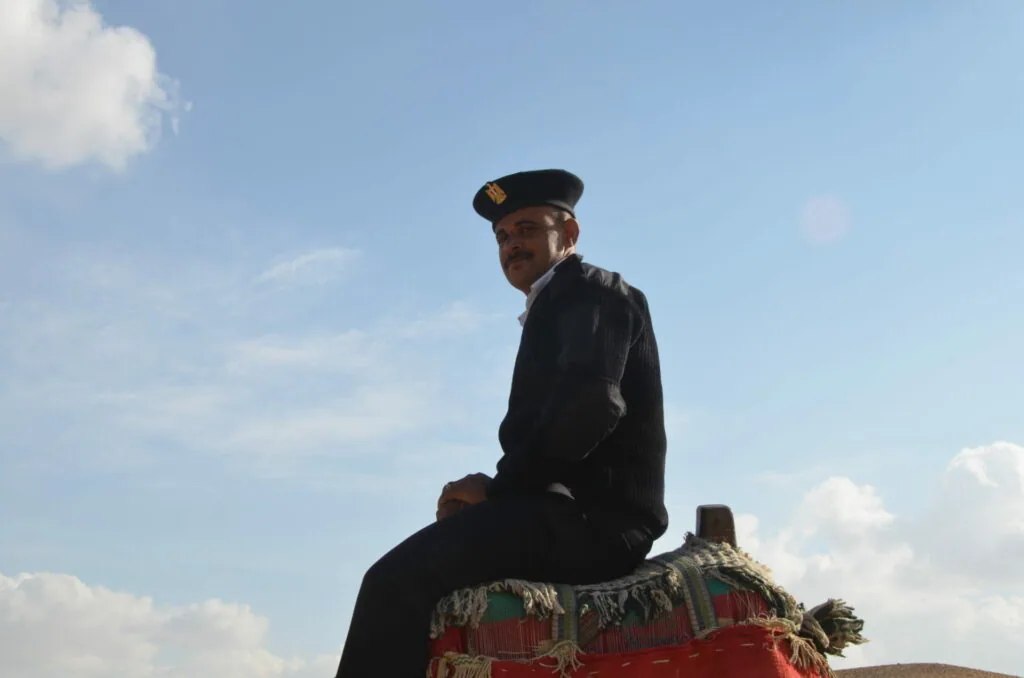 Man in black uniform and cap sitting on decorated red camel against blue sky with clouds