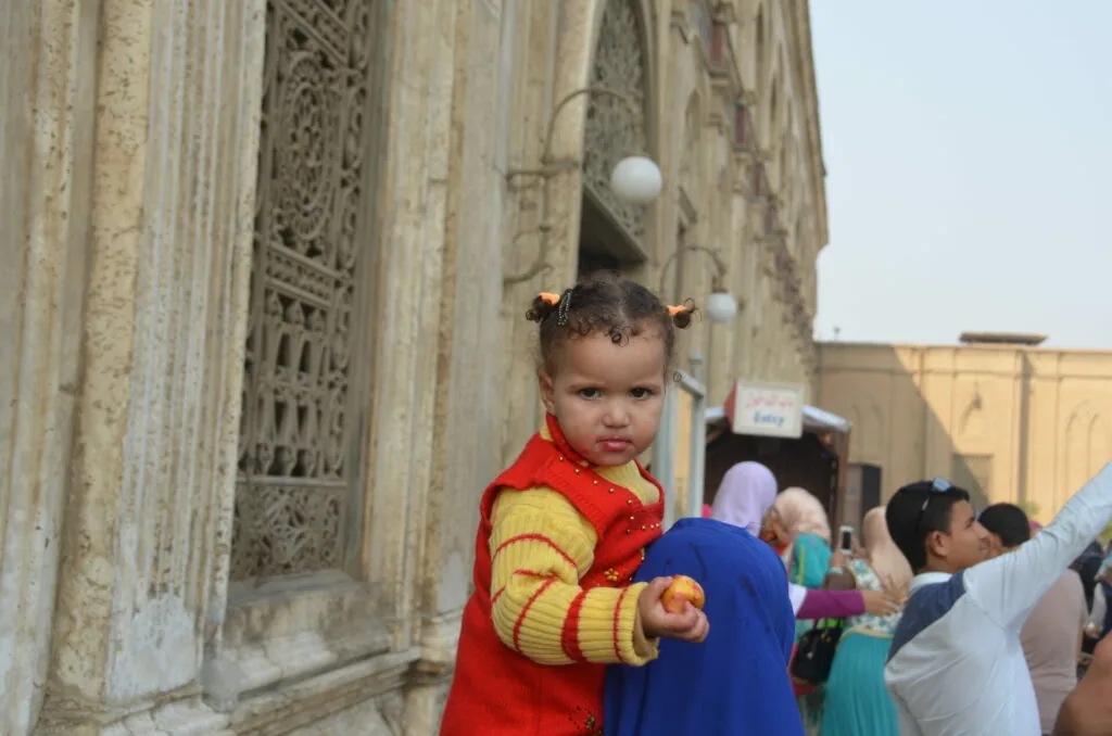 Toddler in red and yellow jacket with hair accessories standing against ornate stone building wall
