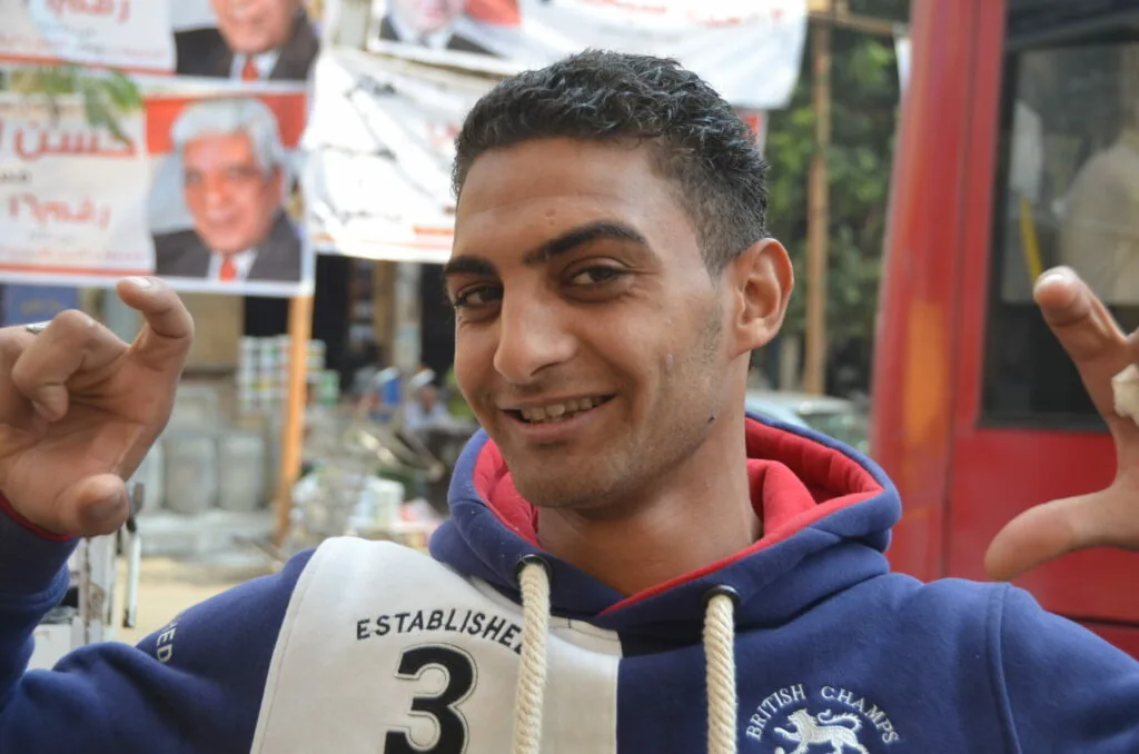 Man in blue and red hoodie smiling with hands raised, outdoor market setting with posters in background