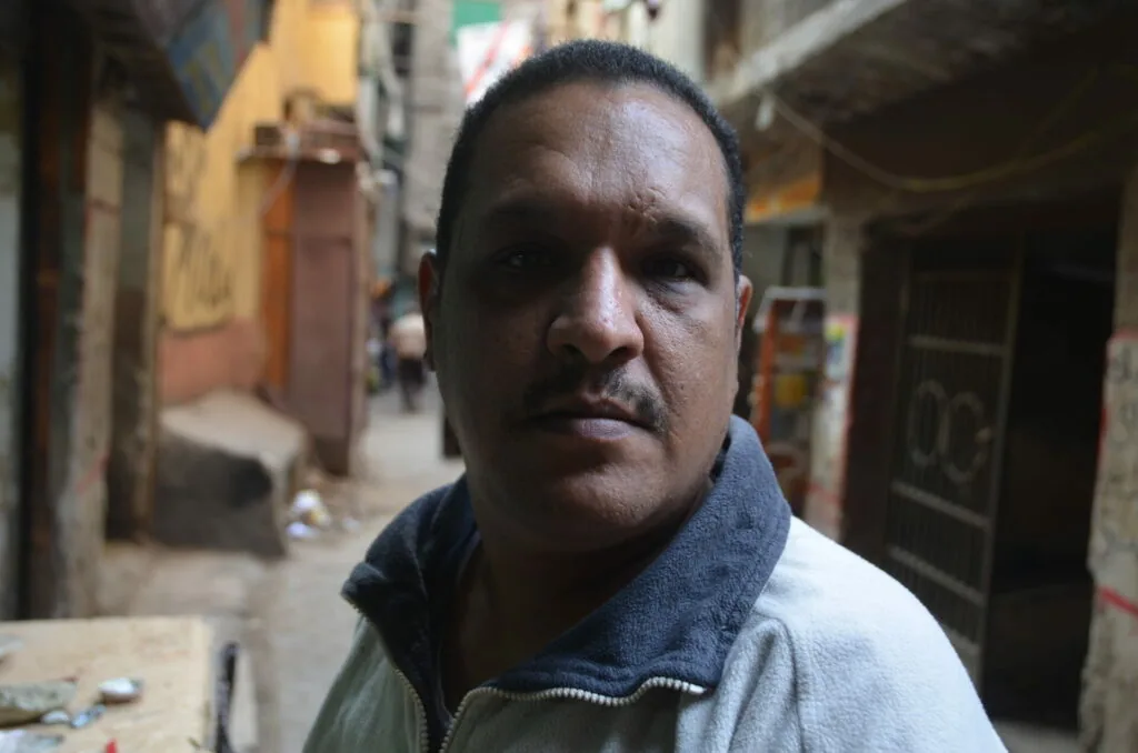 Man with mustache wearing gray and white jacket standing in narrow urban alley with buildings behind him
