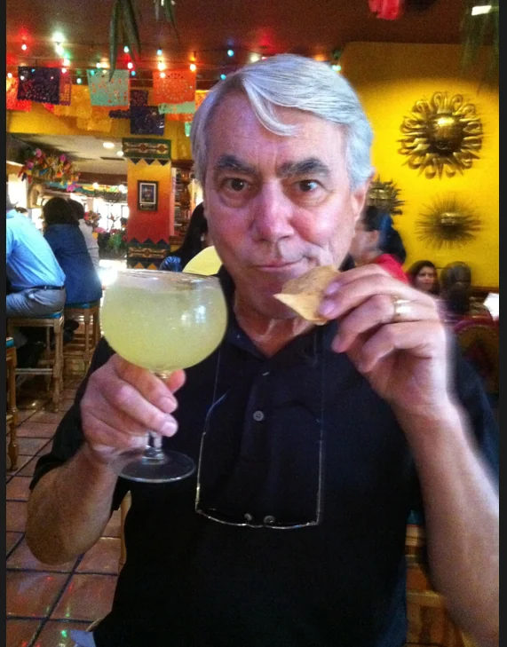 Man with white hair holding a margarita drink in a colorful restaurant setting with decorative sunflowers on yellow wall.