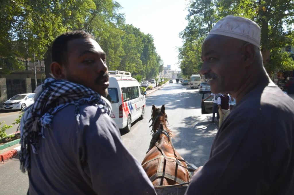 Two men converse on a tree-lined city street with a horse-drawn cart and vehicles visible in the background