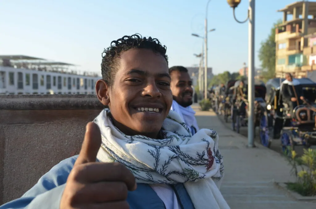 Young man smiling with thumbs up gesture on a waterfront street with buildings and people in background