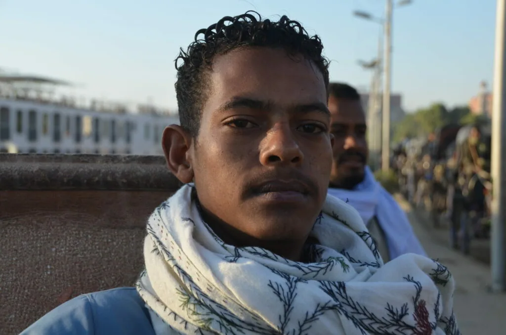Man with dark hair and mustache wearing light blue patterned scarf, standing outdoors with historic white building in background