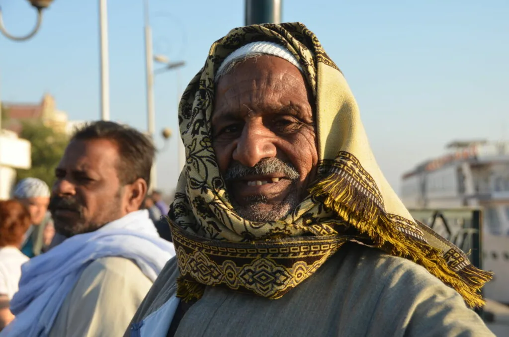 Man wearing white and gold patterned headscarf outdoors with other people in background