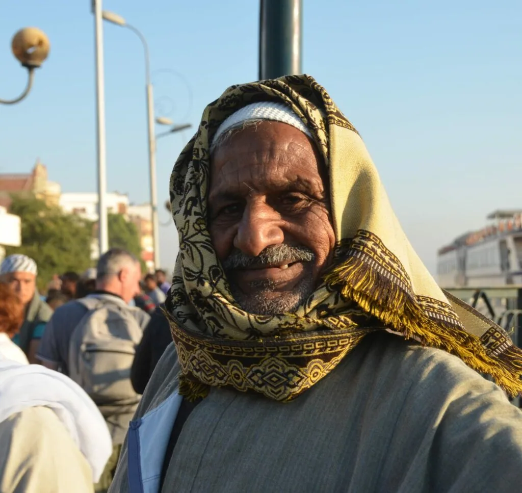 Man wearing a yellow and cream colored head wrap with ornate gold embroidery in an outdoor urban setting