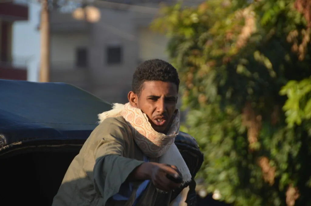 Man in tan and black jacket with white scarf looking over shoulder outdoors