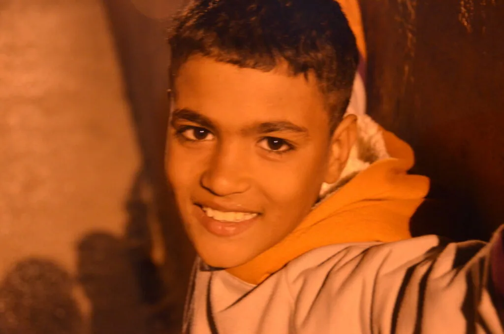 Young person smiling at camera wearing orange and tan jacket against warm-toned background