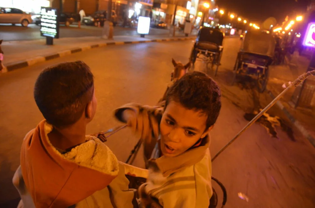 Two young boys playing with a kite on a nighttime street lit by golden streetlights and storefront signs.