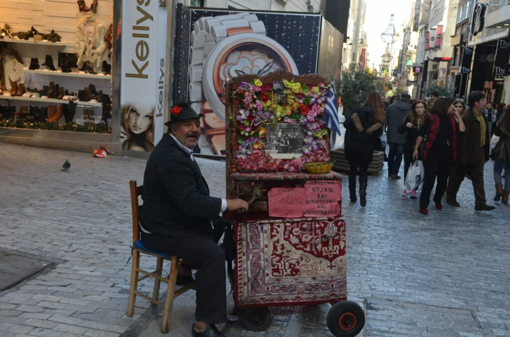 Man in suit sitting on chair beside decorative floral display on wheeled cart in busy pedestrian street
