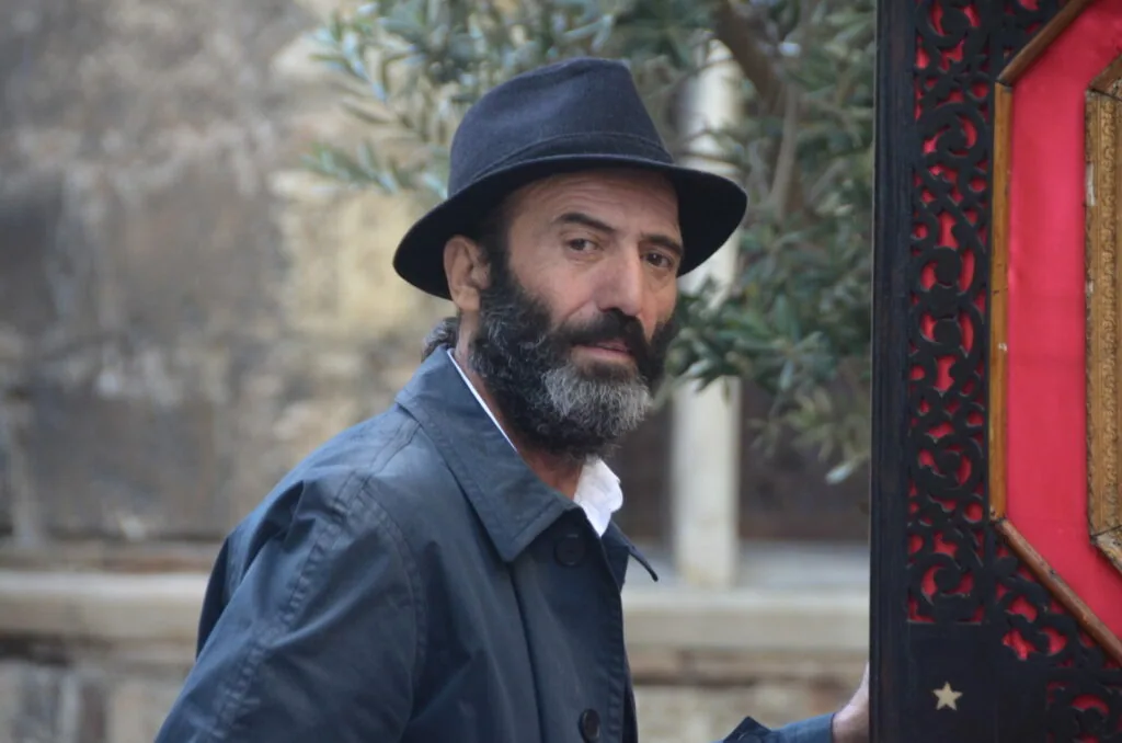 Man with dark beard wearing navy blue fedora hat and shirt standing near red ornate door frame with ivy vines behind him