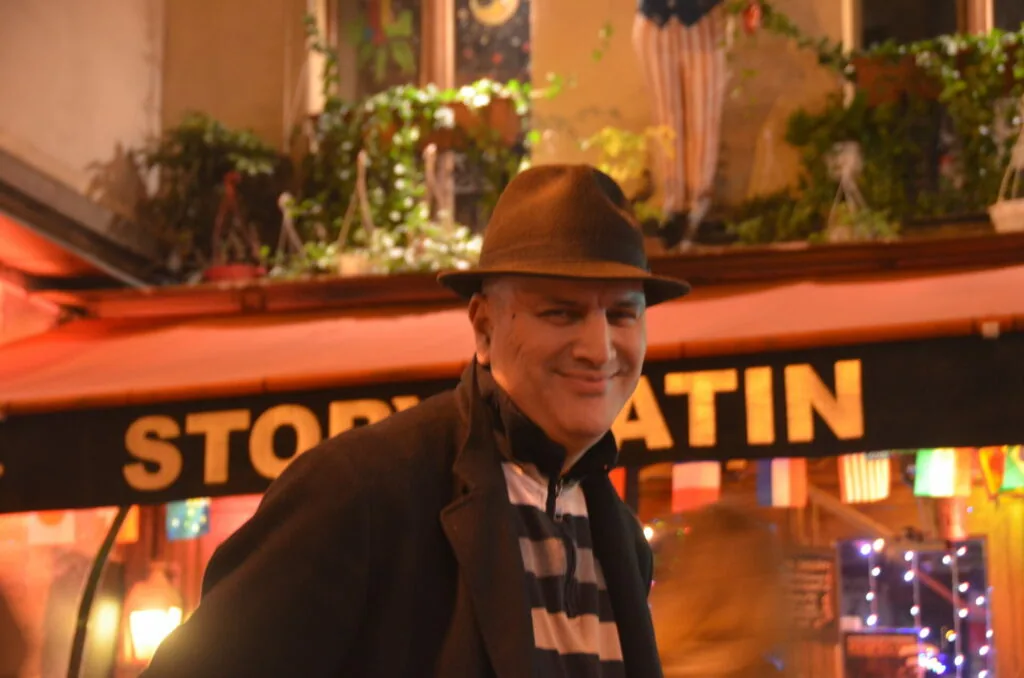 Smiling man wearing brown fedora and dark jacket standing in front of illuminated storefront with hanging plants at night