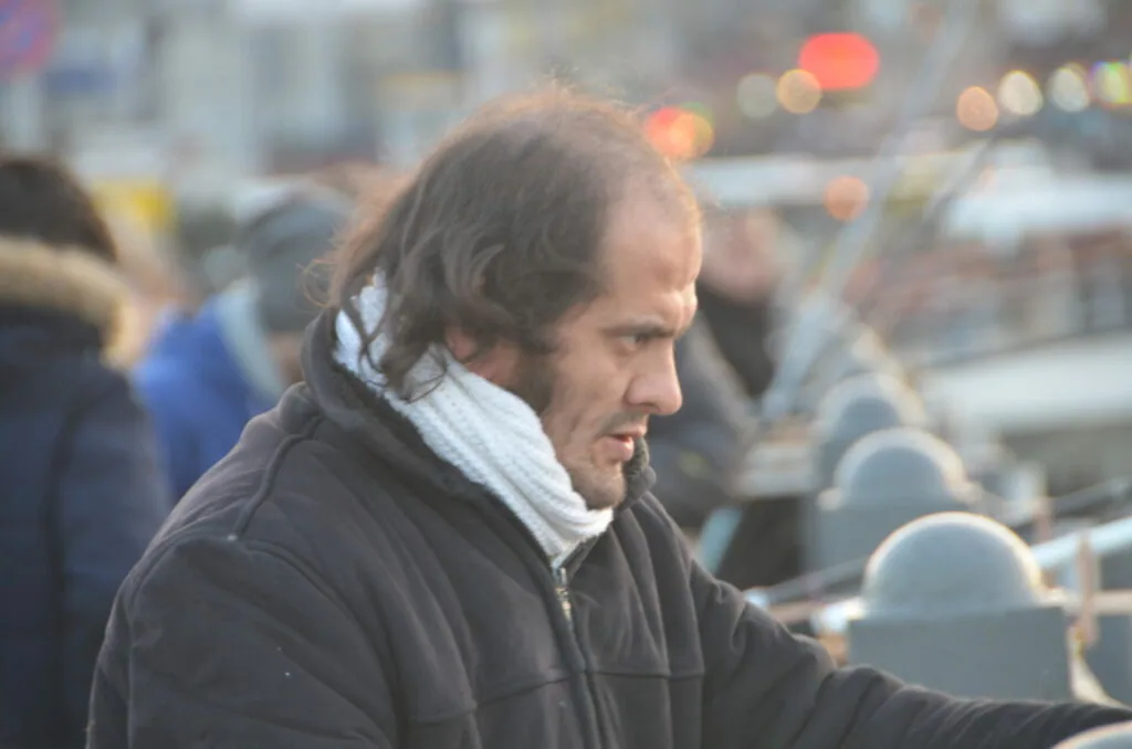Man wearing black jacket and white scarf at crowded outdoor venue with blurred cityscape background