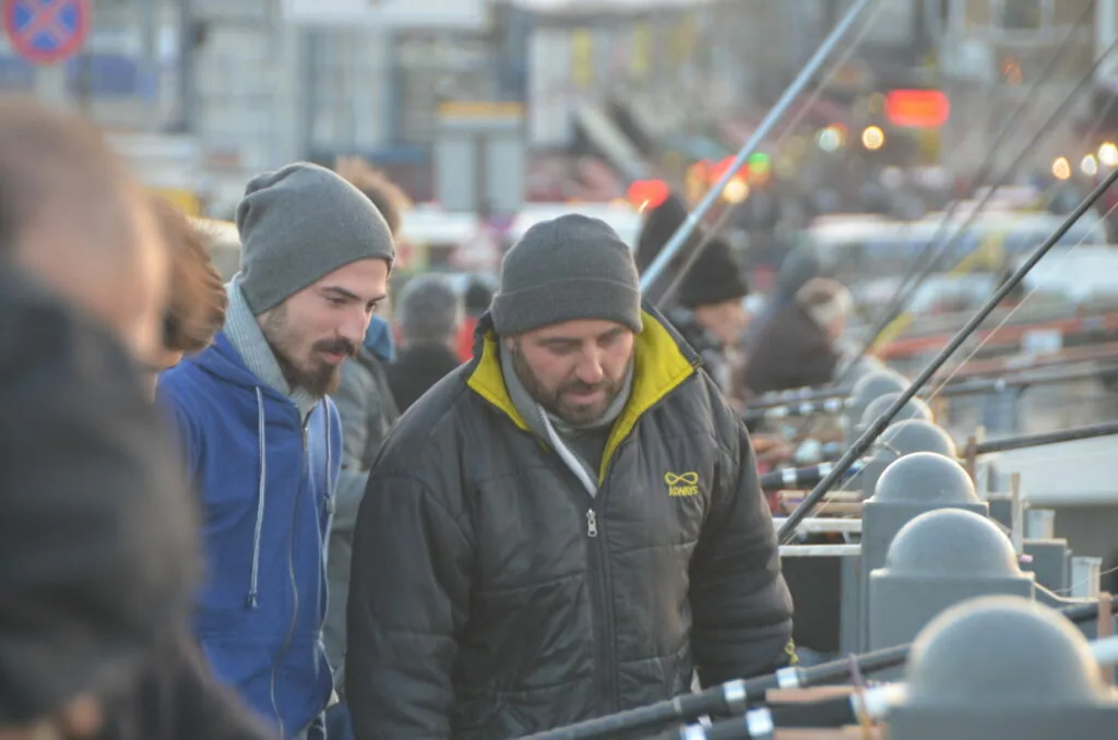 Two men in winter hats and jackets standing on a rooftop overlooking a city street with buildings and traffic lights in the background
