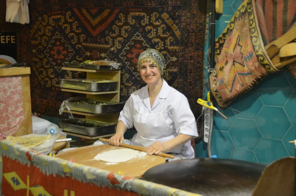 Woman in white chef's coat working at wooden table with stacked metal trays and traditional woven textiles in background