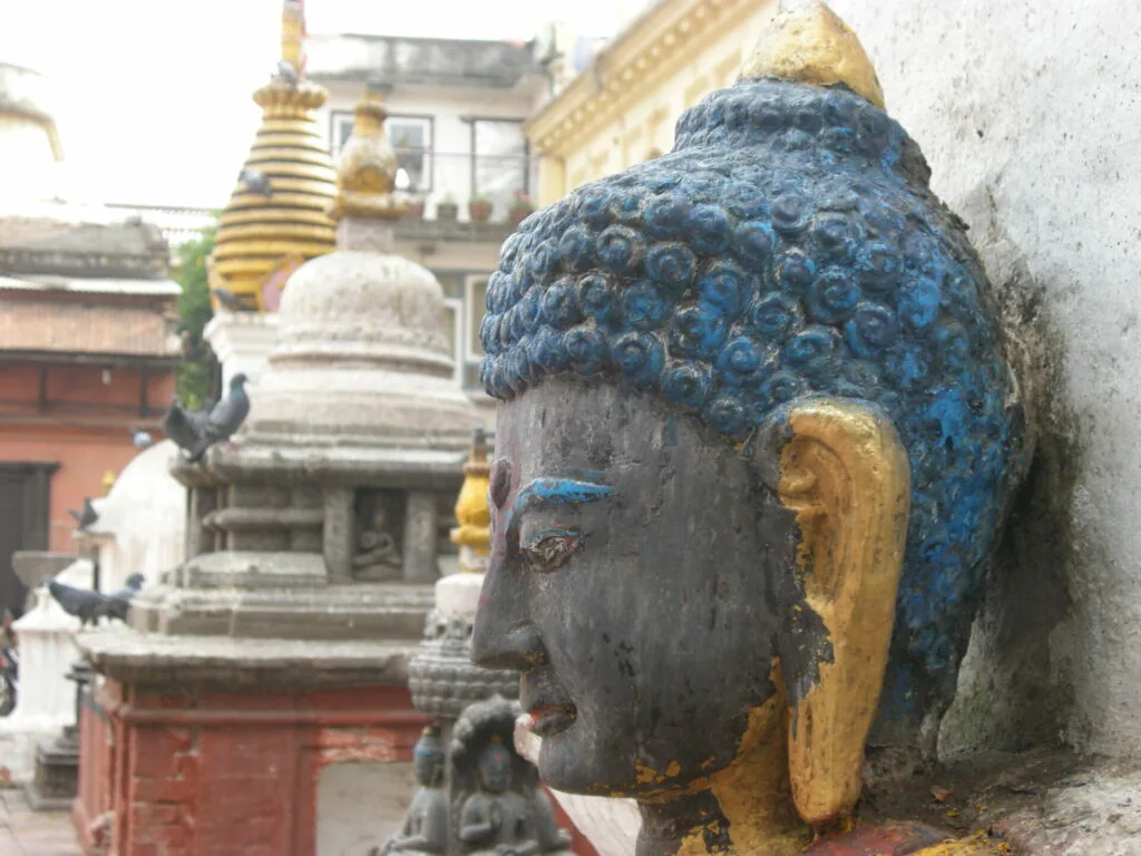 Blue and gold painted stone Buddha head sculpture with golden ear, white and gold stupas visible in background at temple courtyard