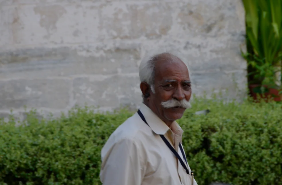 Older man with white hair and mustache wearing cream-colored shirt standing against weathered stone wall with green plants