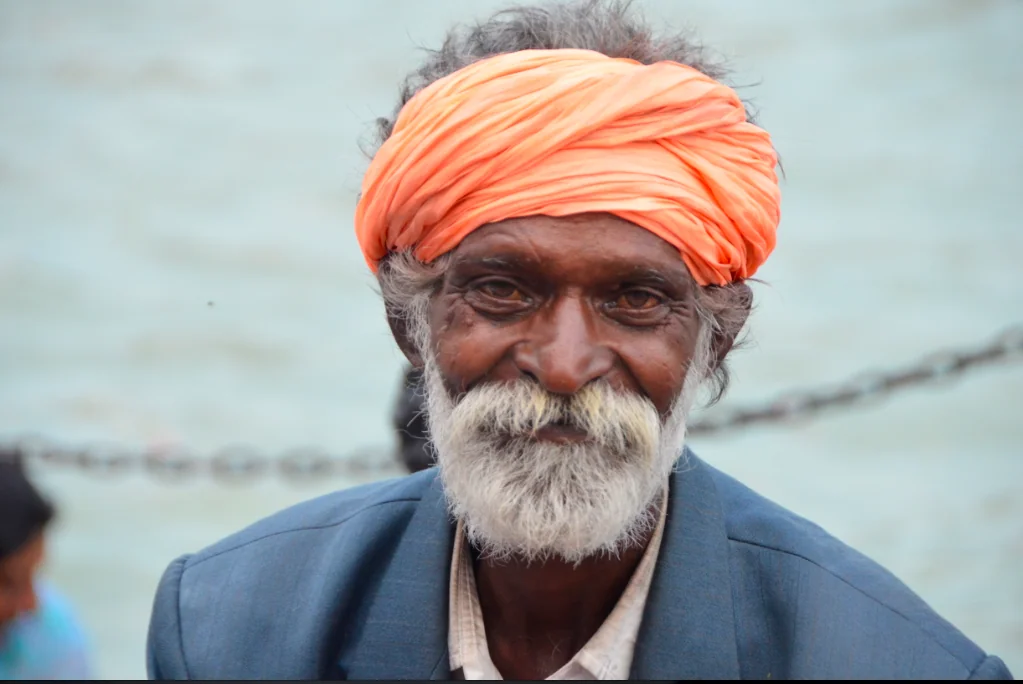 Elderly man with white beard wearing bright orange turban and blue jacket against waterfront background