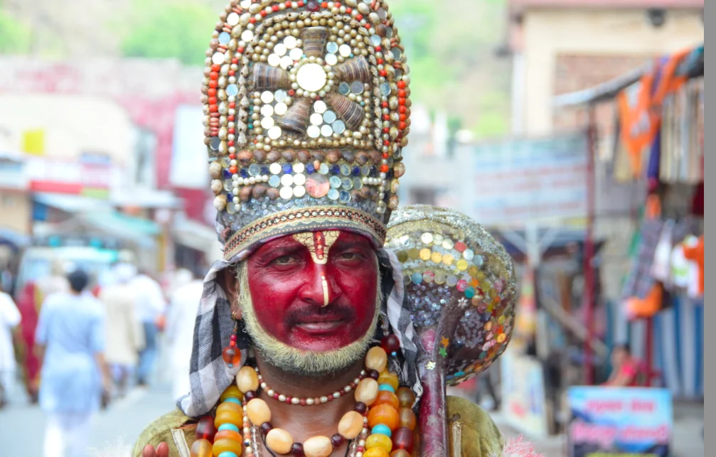 Person wearing elaborate red face paint, ornate beaded headdress with geometric patterns, and colorful beaded necklaces at a crowded street festival or celebration.