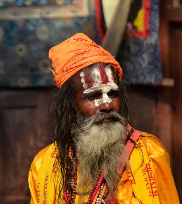 Man with long gray beard wearing orange turban and yellow robe with red markings and beaded necklaces