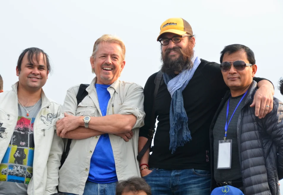 Four men standing together outdoors smiling at camera, wearing casual clothing and accessories including a yellow cap and sunglasses