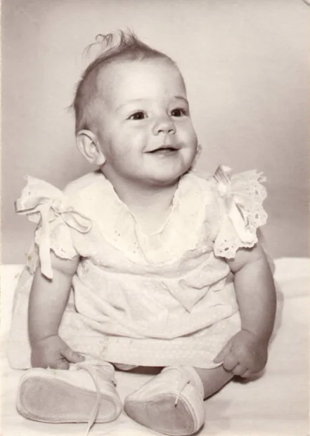 Smiling infant in white ruffled dress sitting against neutral background