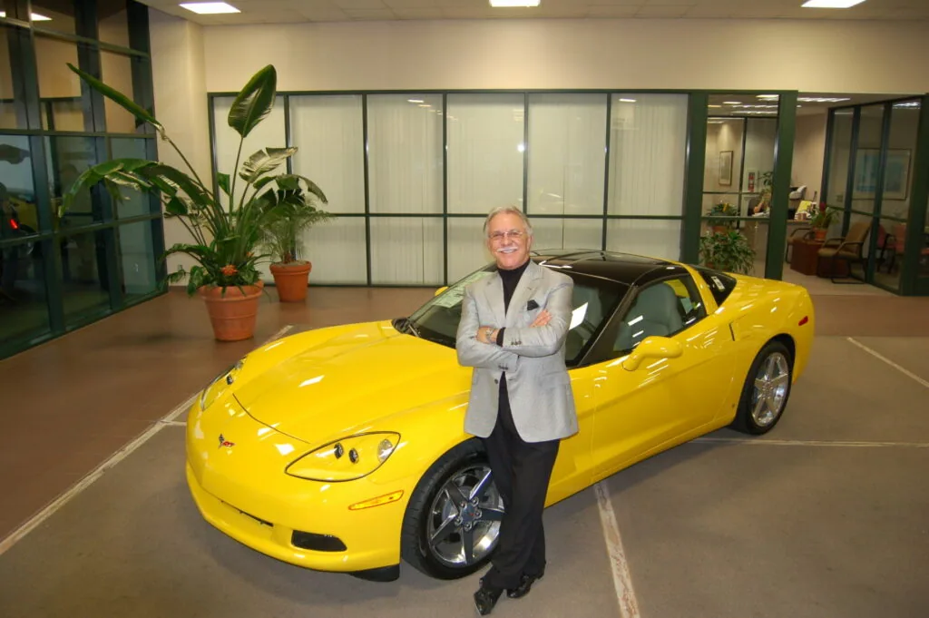 Man in light gray blazer standing next to bright yellow sports car in modern showroom with potted plants and glass partitions