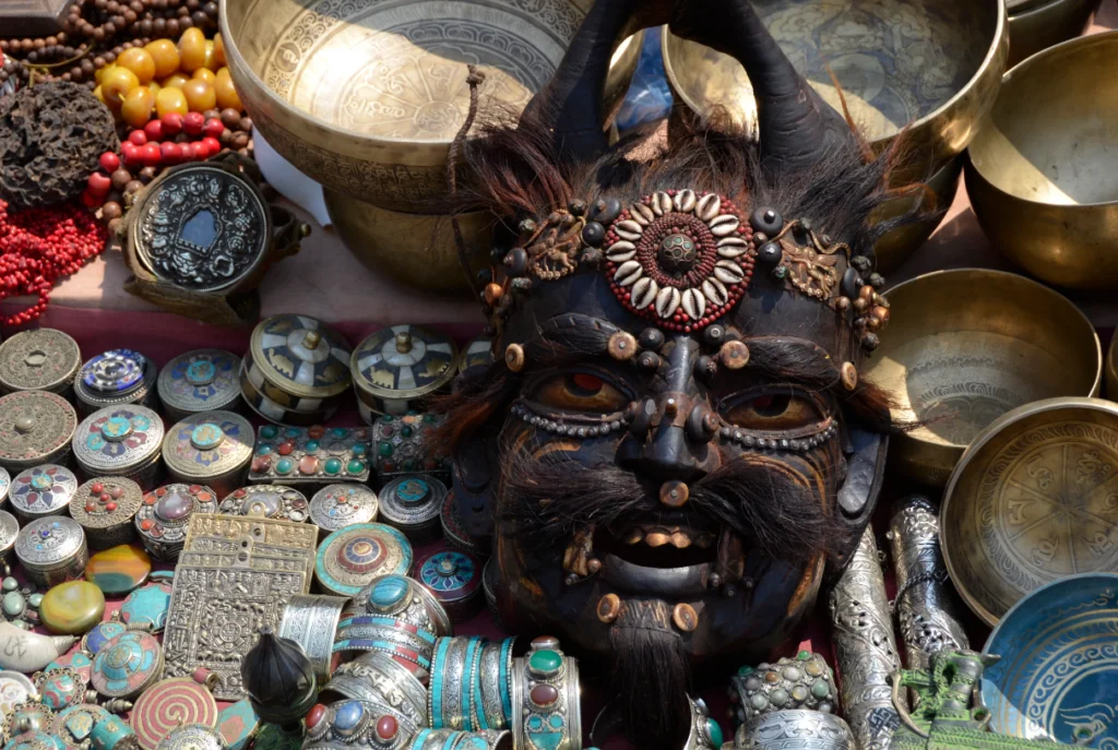 Black wooden demon mask with horns surrounded by brass singing bowls, beaded necklaces, decorative plates, and spiritual artifacts at an international market display