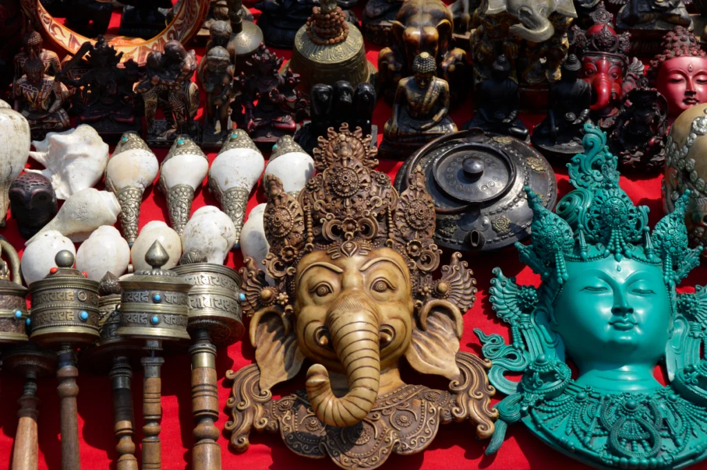 Collection of ornamental figurines displayed on red cloth including white cone-shaped objects, brass deity masks, metal prayer wheels, and turquoise carved sculptures
